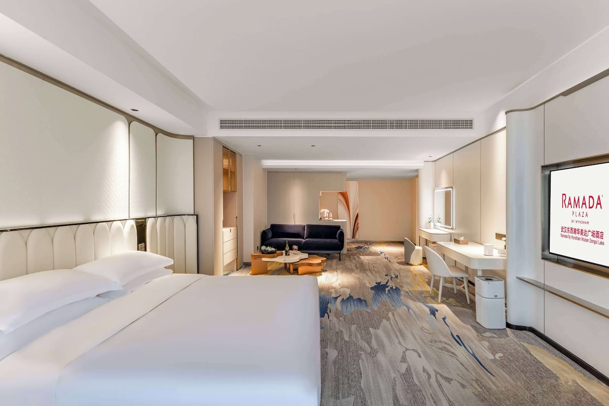 A modern hotel room with a large bed, a sofa, a desk, and a television displaying the hotel name.