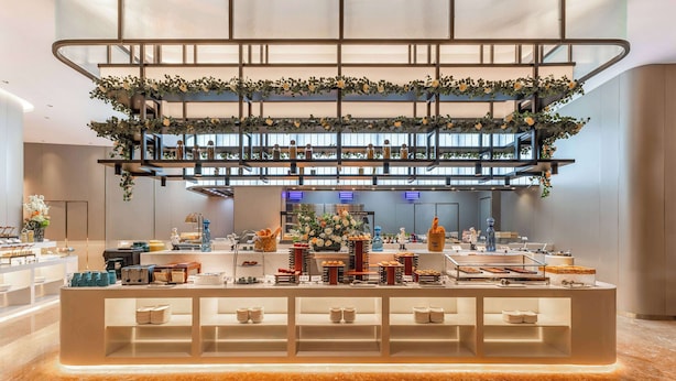 A modern hotel lobby with a central reception desk, a staircase with greenery, and a view of the kitchen area. - Ramada Plaza By Wyndham Wuhan Dongxi Lake (Wuhan)