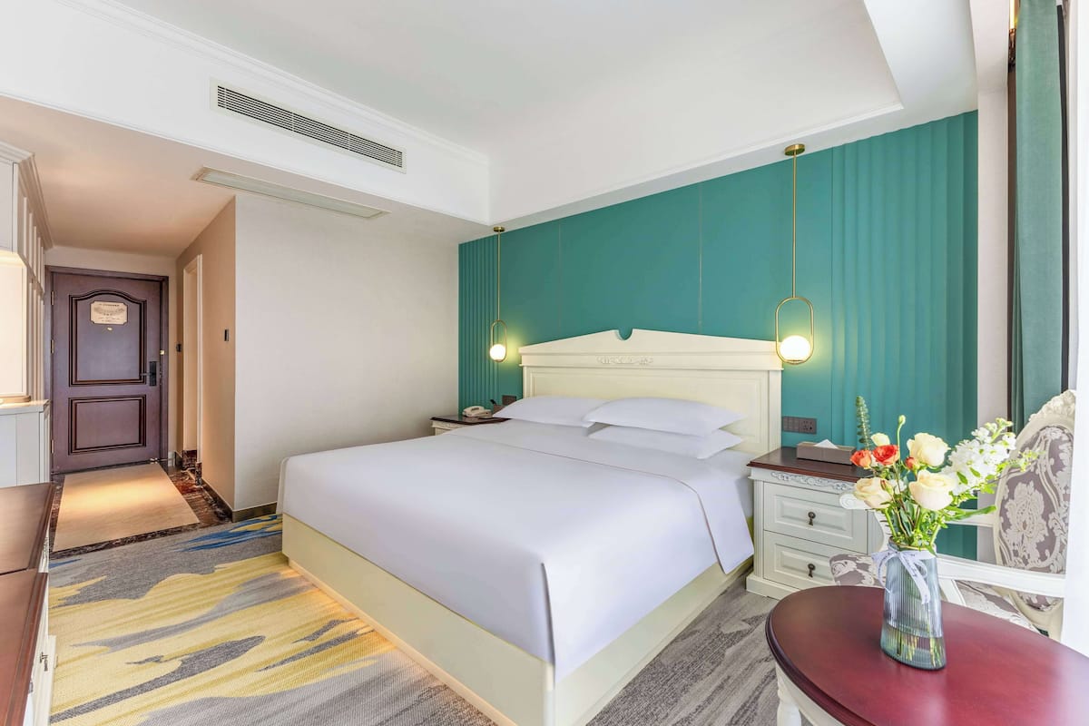 A hotel room with a bed, bedside table, nightstand, and a vase of flowers.
