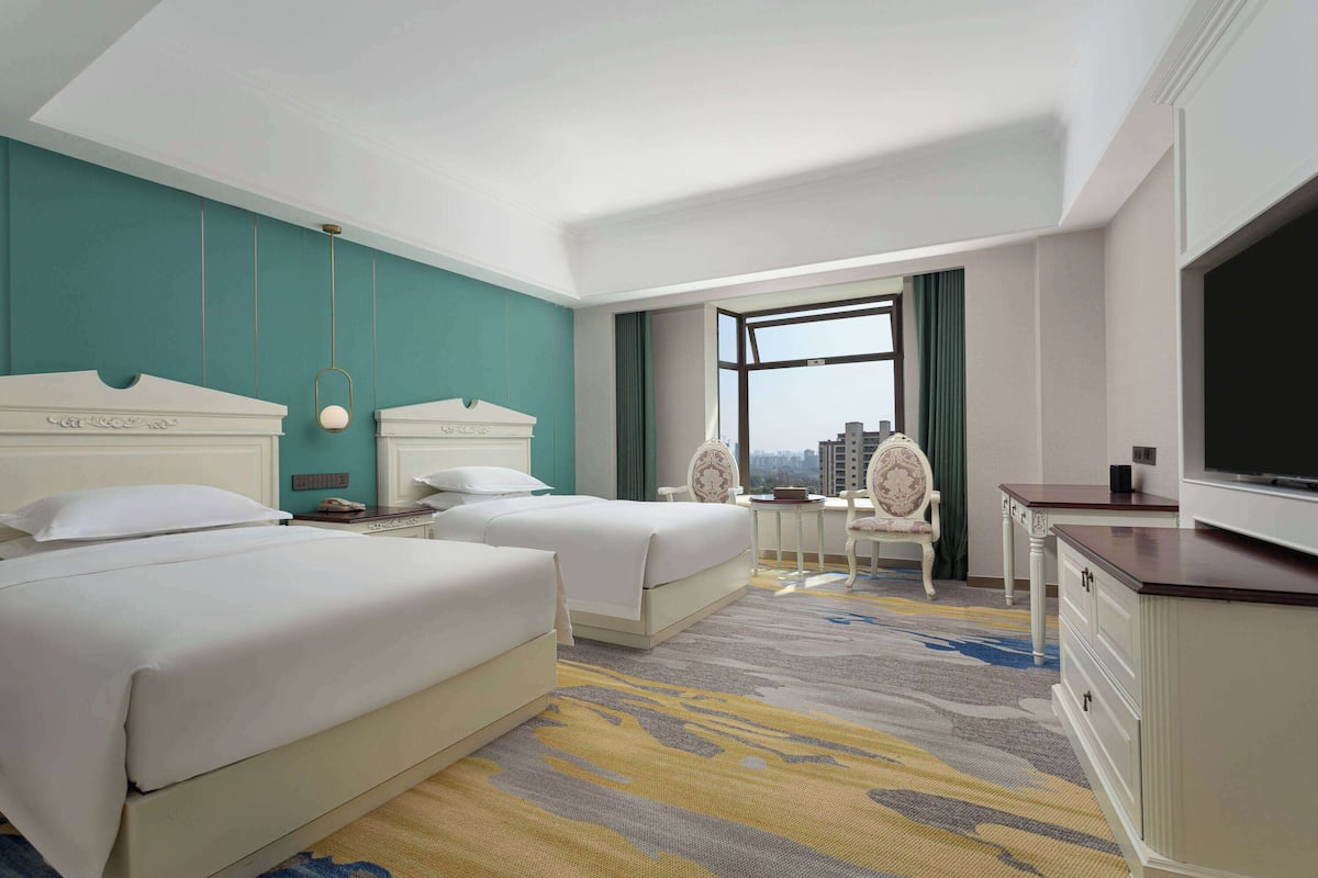 A hotel room with two beds, a desk, a chair, a TV, and a view of the city.