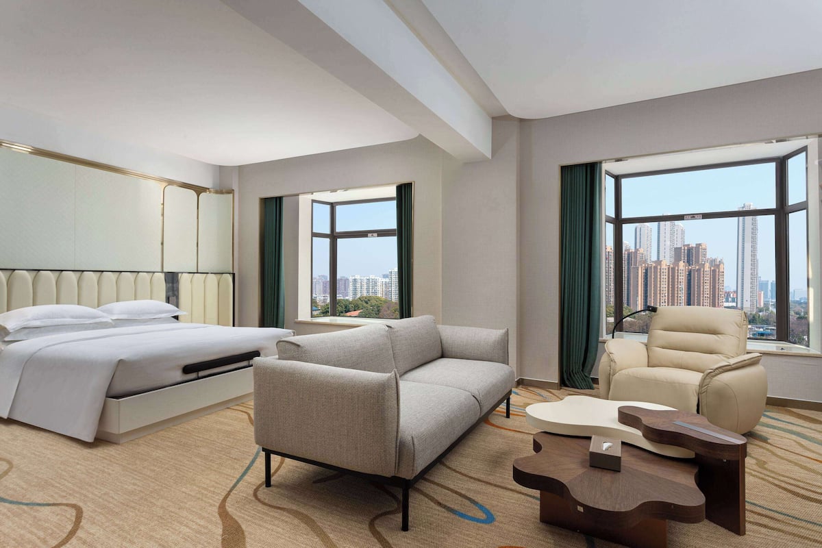 A modern hotel room with a bed, sofa, armchair, and a view of the city skyline.