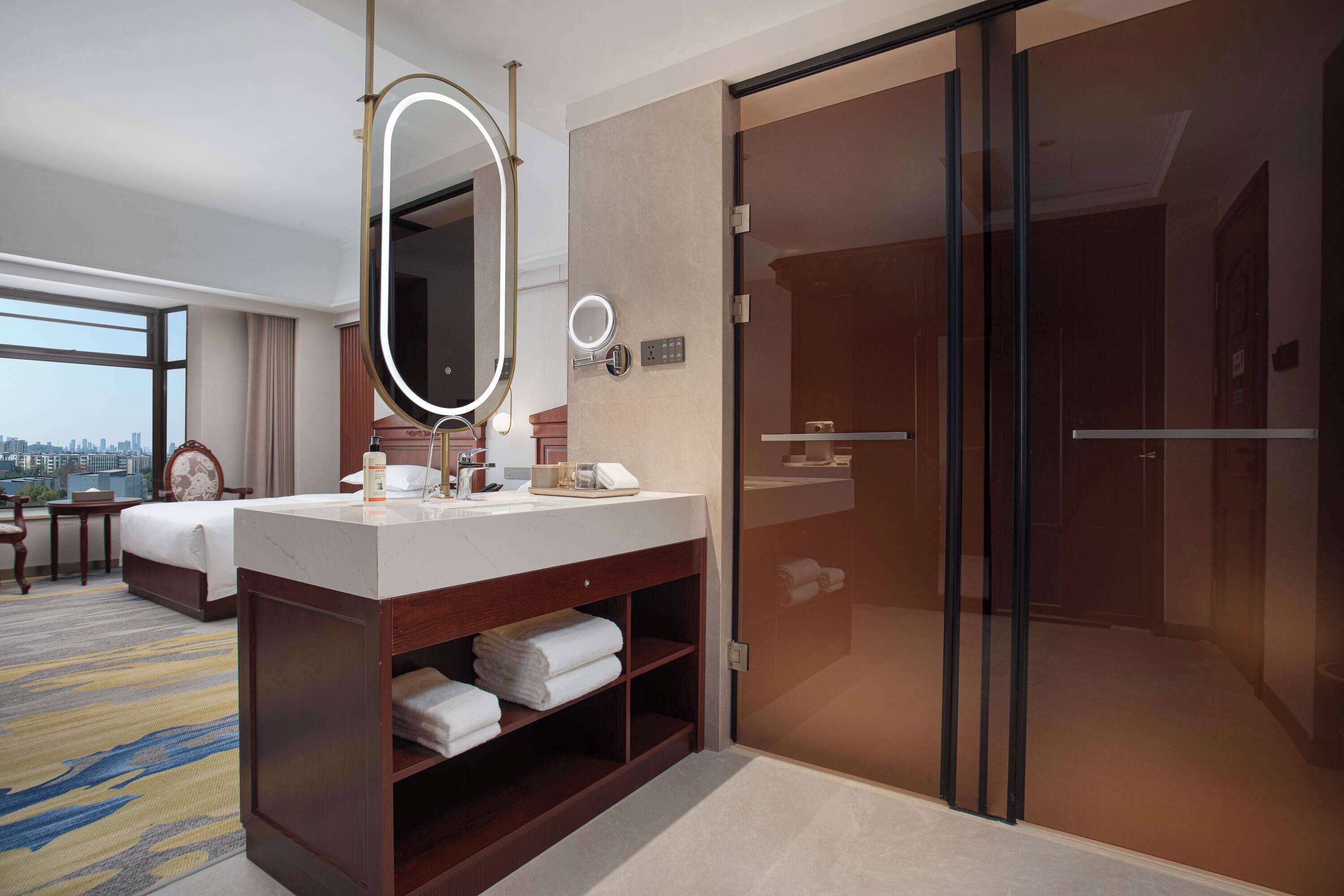 A hotel room with a large bed, a vanity with a mirror, a shower, and a view of the city.