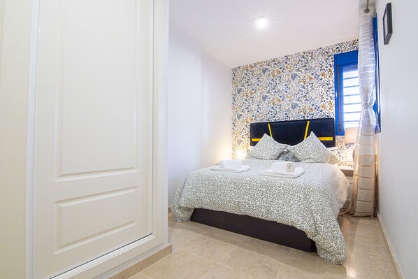 Ein Schlafzimmer mit Bett, Nachttisch, Fenster mit Vorhängen und einer Wand mit einem Blumenmuster.