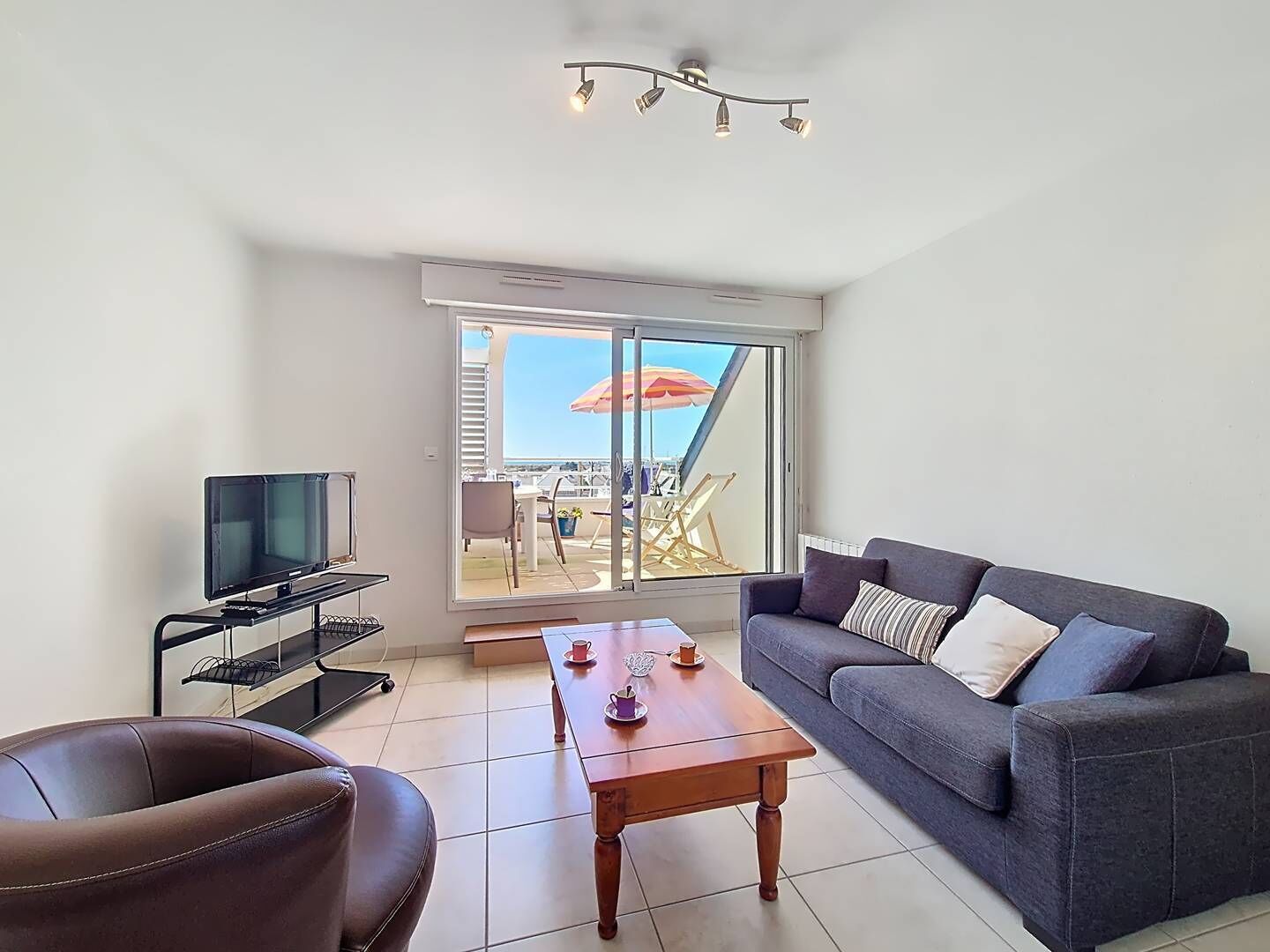 Ein Wohnzimmer mit einer Couch, einem Couchtisch, einem Fernseher auf einem Schrank und einem Balkon mit Gartenmöbeln.