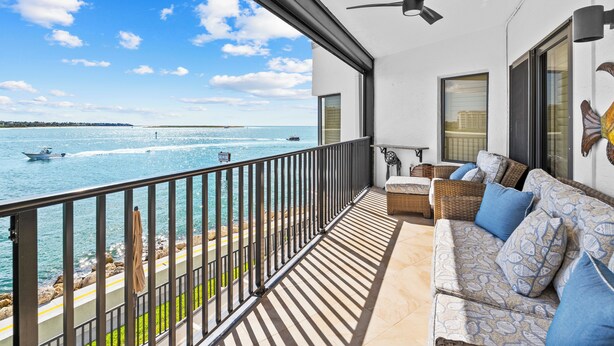 A balcony with a view of the sea, a sofa with patterned cushions, a ceiling fan, and a window with blinds.