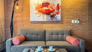 Un salon moderne avec un canapé gris, deux coussins rouges, une table basse sur laquelle sont disposées des pâtisseries, une tasse de café et un tableau accroché au mur de briques.