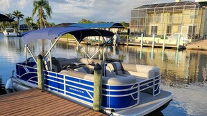 A pontoon boat docked at a wooden pier with a covered canopy, a house with a balcony in the background, and a calm water body reflecting the sky.