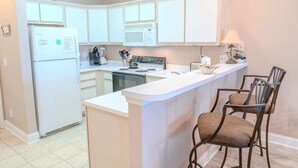 Een keuken met witte kastjes, een fornuis, een magnetron, een koelkast en een bar met twee stoelen.