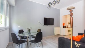 Una sala de estar moderna con una mesa de comedor de vidrio, sillas negras, un televisor de pantalla plana montado en la pared y una lámpara de pie decorativa.
