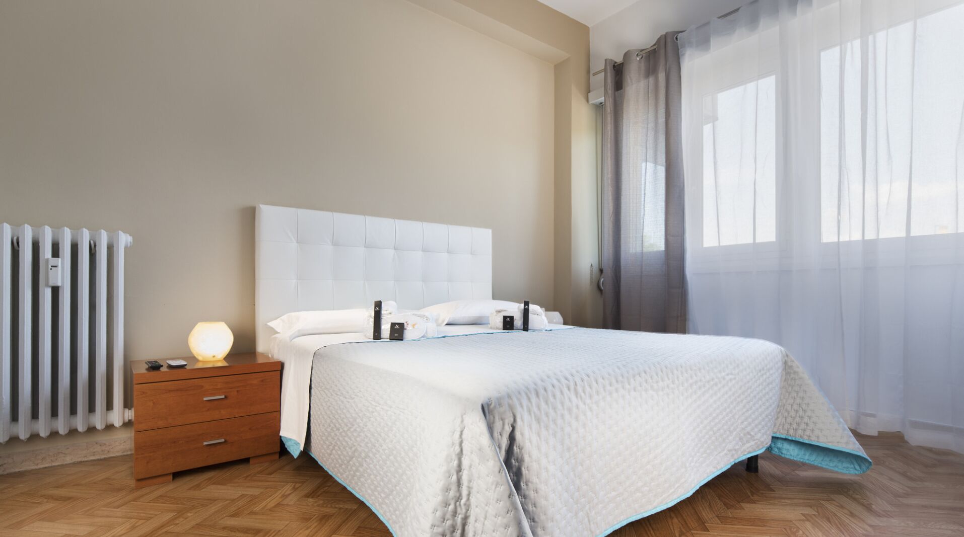 A modern hotel room with a large bed, a wooden nightstand with a lamp, a radiator, and sheer curtains.