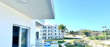 A modern building with a balcony, a pool, and a view of other buildings and greenery.