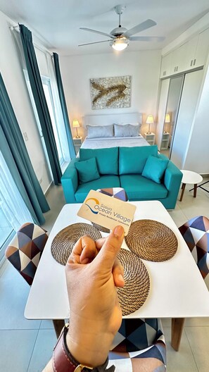 Una habitación de hotel con una cama, un sofá color turquesa, una mesa con individuales tejidos y una mano señalando un folleto.