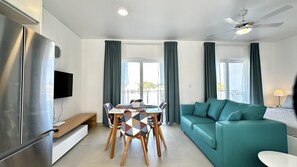 Ein modernes Wohnzimmer mit einem türkisfarbenen Sofa, einem Esstisch mit gemusterten Stühlen, einem Flachbildfernseher und einem Deckenventilator.