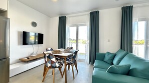 Ein modernes Wohnzimmer mit einem türkisfarbenen Sofa, einem Esstisch mit Stühlen, einem flachbildfernseher an der Wand und einem Balkon mit Vorhängen.