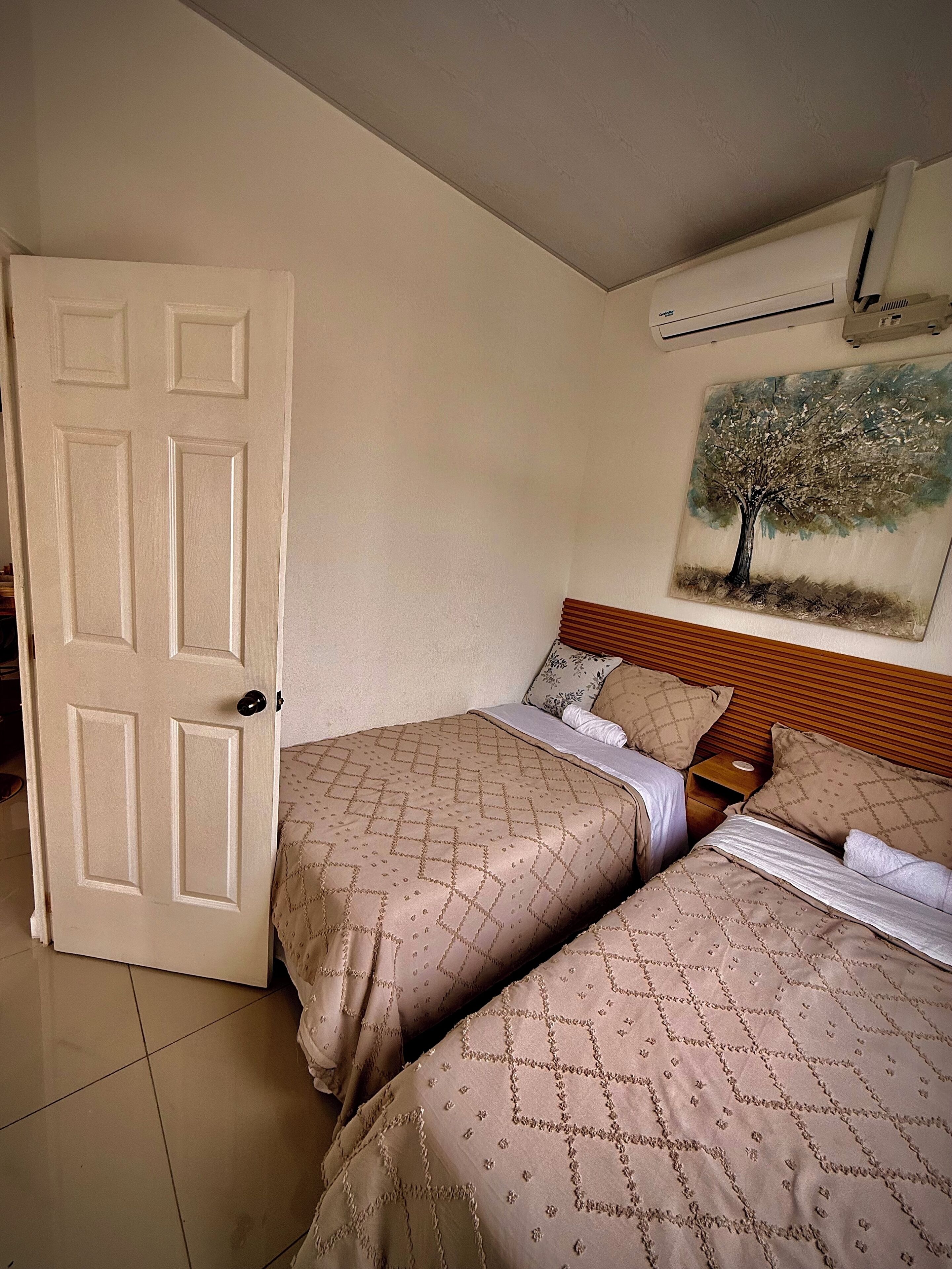Un dormitorio con dos camas, una puerta, un cuadro de un árbol y un equipo de aire acondicionado.