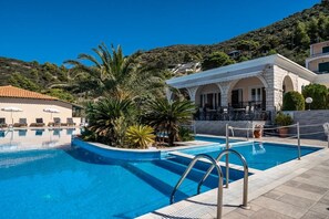 Una piscina con solárium de piedra, palmeras y un edificio con mesas al aire libre.