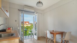 An open kitchen with a dining area, a sliding glass door to a balcony, and a view of a house. - Apartment '' with Shared Garden, Wi-Fi and Air Conditioning (Póros Kefalonias)