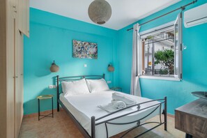 Une pièce comprenant un lit, une table de chevet, un luminaire suspendu, une fenêtre avec des rideaux et une œuvre d’art accrochée au mur.