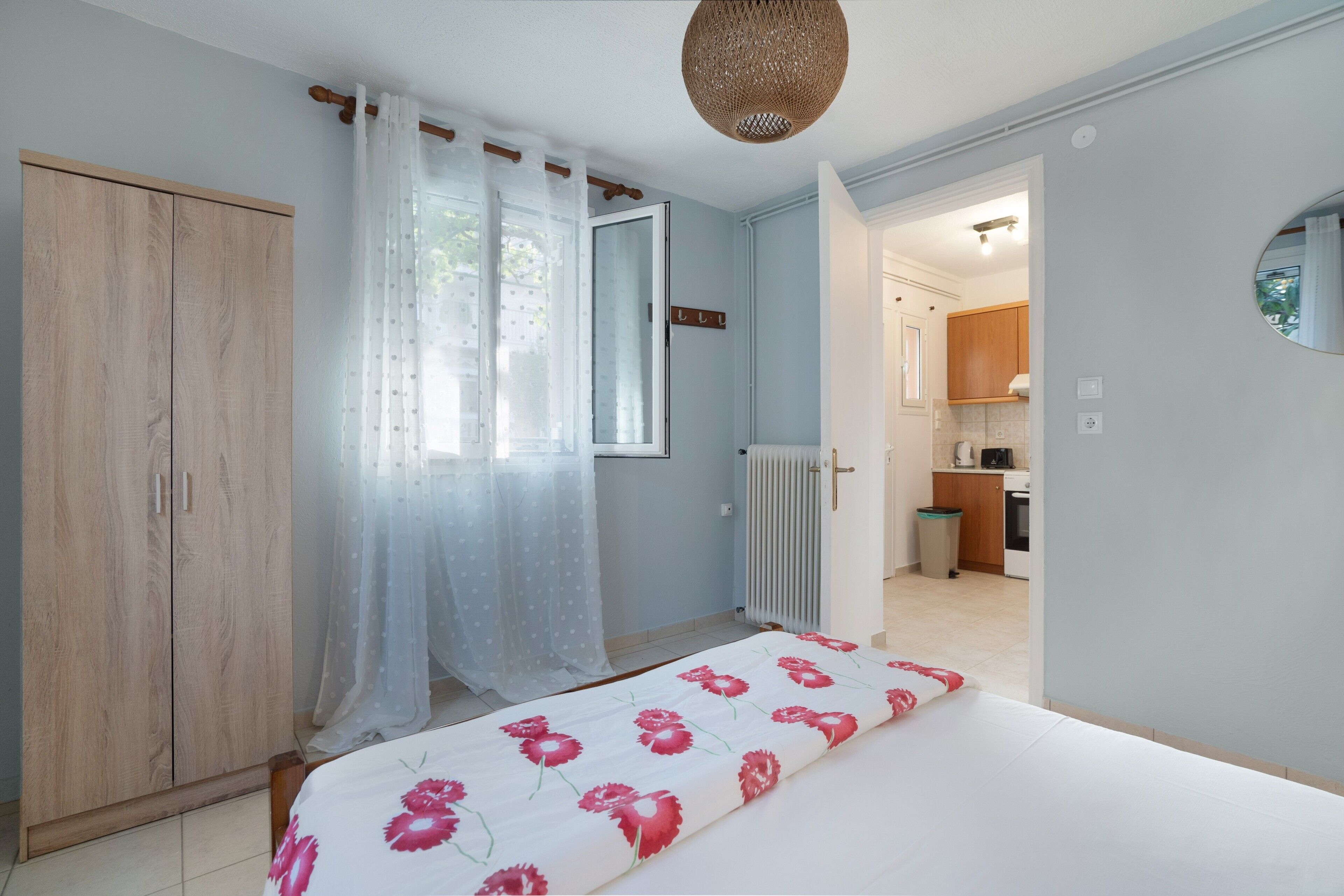 Un dormitorio con cama, armario, ventana con cortinas translúcidas y puerta abierta que da a una cocina.