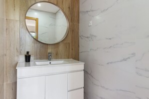 Une salle de bain avec un mur en marbre, un panneau en bois, un meuble-lavabo blanc avec un évier et un miroir rond.