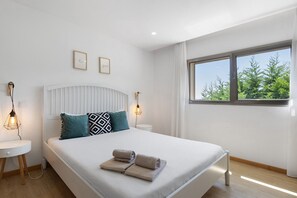 A modern hotel room with a large bed, two bedside tables, a window with a view of trees, and two wall-mounted light fixtures.