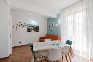 A dining area with a table, chairs, a sofa, and a window with curtains. - Apartment 'Kairos' with Shared Pool, Wi-Fi and Air Conditioning (San Vito Lo Capo)