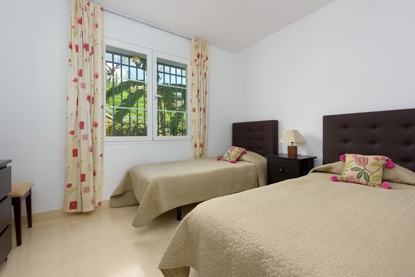 A room with two beds, a window with curtains, a bedside table with a lamp, and a view of greenery outside.