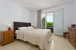 Une chambre à coucher comprenant un lit, des tables de chevet, une commode et une porte-fenêtre coulissante donnant sur un balcon.