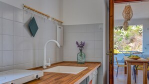 Een keuken met een houten aanrecht, witte tegels, een witte spoelbak, een afwassigheid, een glazen vaas met bloemen en uitzicht op het buiten.