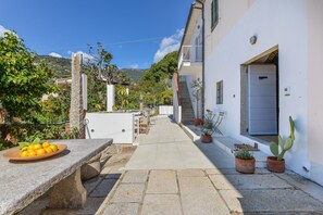 A courtyard with a stone bench, a table with a fruit bowl, potted plants, and a staircase leading to a white building.