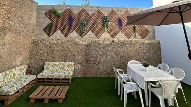 Une terrasse extérieure avec un mur en pierre, du gazon synthétique, des meubles blancs et une décoration murale à motifs.