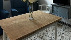 Une pièce avec une table en bois, une chaise bleue, un vase avec des fleurs, un téléviseur et une fenêtre avec des stores.