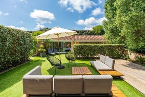Un patio con sofá, sillas y una mesa, una casa pequeña con techo de tejas y un jardín cuidado.