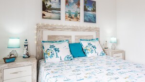 Una cama con cubrecama de temática náutica, un cabecero con luces de cadena y dos cuadros colgados en la pared que representan escenas tropicales.