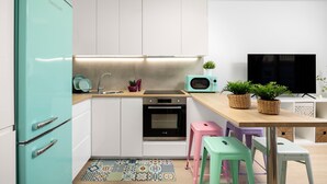 Una cocina compacta con un refrigerador verde azulado, armarios blancos, un microondas y un horno pequeño. Hay dos taburetes y un televisor en la pared.