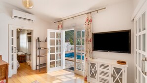 A room with a wooden floor, a white cabinet with glass doors, a flat-screen TV mounted on the wall, and a door leading to an outdoor area with a pool. - Vacation 'Pool House' with Mountain View, Wi-Fi and Air Conditioning (Vega de San Mateo)
