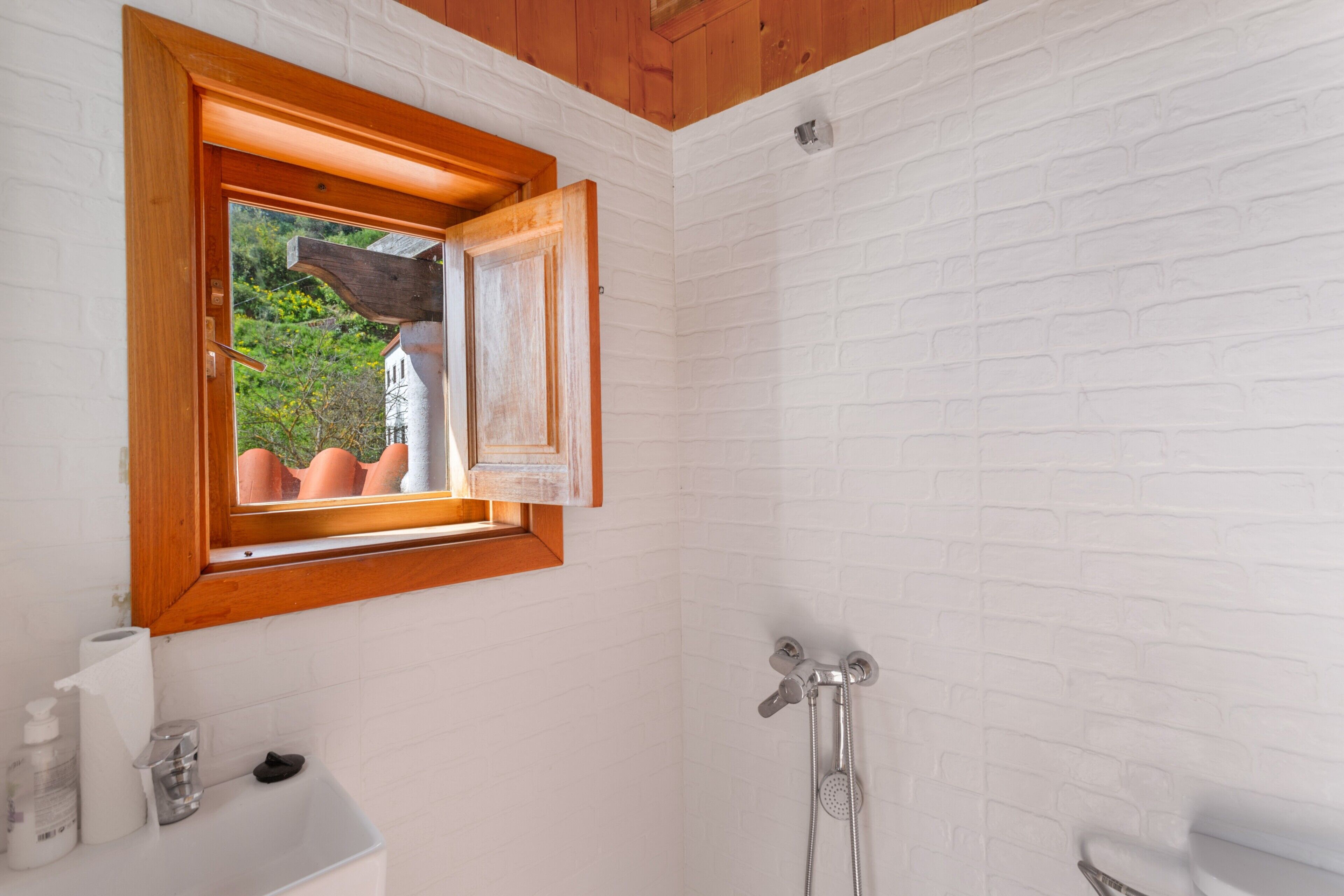 A bathroom with a white brick wall, a wooden-framed window, a shower, and a sink with toiletries.
