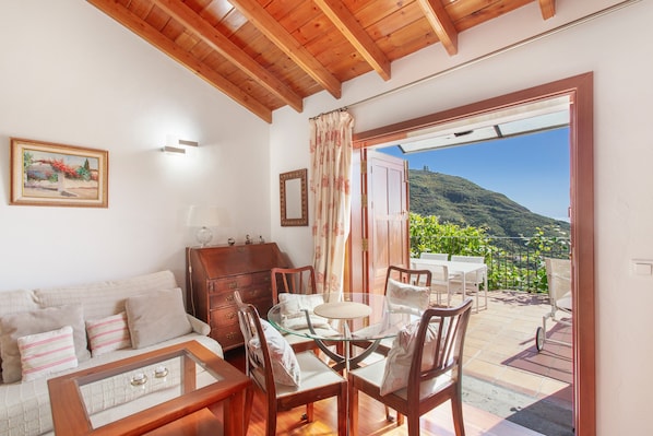 A cozy room with a sofa, wooden cabinet, and a glass table. It features a view of a mountain and a balcony with outdoor furniture.