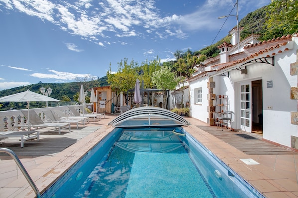 A swimming pool with a sunshade, lounge chairs, and a view of a hillside.