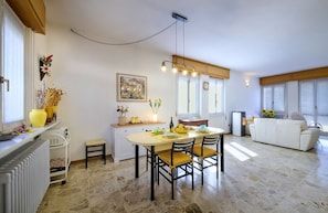Un espace repas comprenant une table, des chaises, un radiateur, un petit tabouret, une étagère fixée au mur avec des objets décoratifs et un luminaire suspendu.