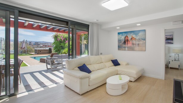 Una sala de estar moderna con un sofá blanco, una mesita redonda y un amplio ventanal con vistas a una piscina y jardín.