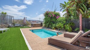 Una piscina con muelle de madera, sillas de descanso y un área de césped con valla.