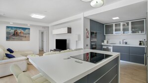 Una cocina moderna con una isla central, un comedor con mesa blanca y una sala de estar con sofá y un cuadro.