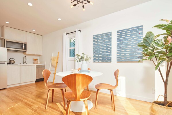 A modern kitchen with white cabinets, a microwave, and a sink. There is a dining area with a white table and wooden chairs.