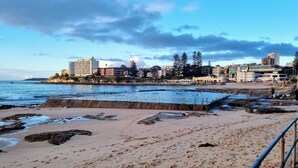 A beach with a metal railing, sandy shore, and buildings in the background. - Cronulla Ocean View, Steps to Beach (Cronulla)