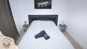 Una cama bien hecha con una colcha blanca y gris estampada, dos toallas enrolladas, un cabecero negro y un cuadro con la palabra 'Hollywood' enmarcado en la pared.