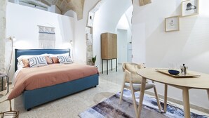 A bedroom with a bed, a dining table, and a chair. There are arches in the ceiling and a wall-mounted artwork.