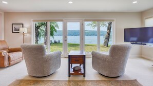 A living room with two armchairs, a small table, a sofa, and a TV. It features large glass doors leading to a lakeside view.