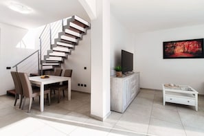 Un salon moderne avec un escalier, une table à manger entourée de chaises, un téléviseur posé sur un meuble et un tableau encadré accroché au mur.