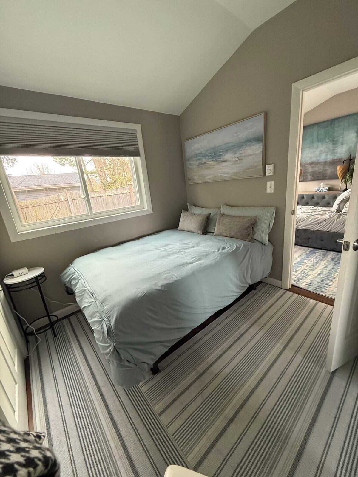 A bedroom with a bed, a window, a painting, and a door leading to another room.